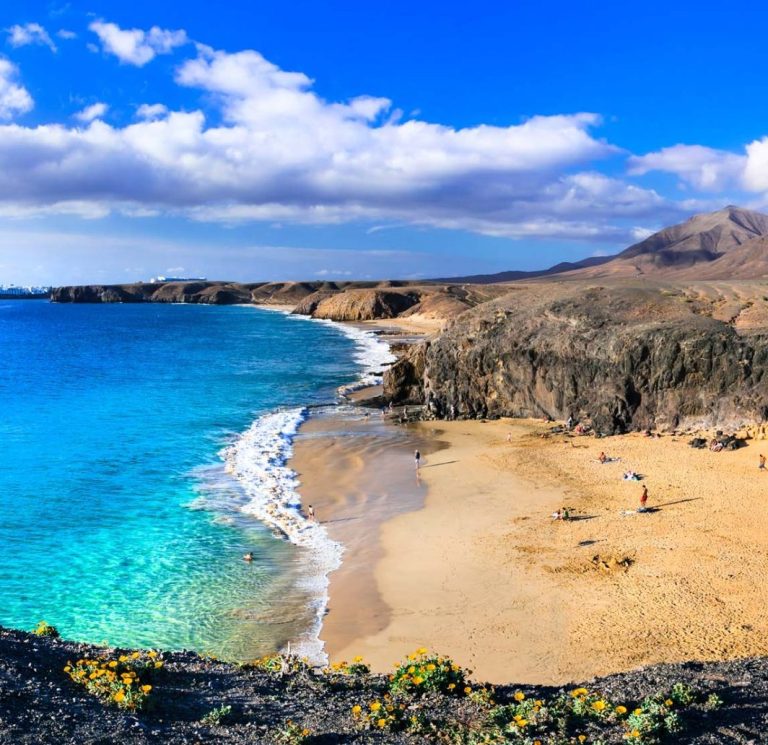 Küstenlandschaft mit klarem Wasser, Sandstrand und Bergen im Hintergrund.